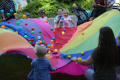 Chusta w barwach tęczy, a na niej podskakujące kolorowe piłki. Brzego chusty trzymają: troje  dzieci i trzy kobiety.