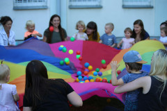 Kolorowa chusta,  a na niej piłki. Brzegi materiału trzyma grupa kobiet i dzieci.