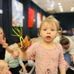 Dziecko trzyma w prawej dłoni doniczkę z roślinką. Patrzy w obiektyw. Różowa koszulka pobrudzona ziemią. W tle uczestnicy zajęć. 