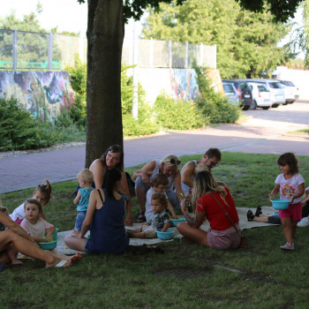 Na trawie pod drzewem siedzą dorośli i dzieci. W tle chodnik, murek i zaparkowane samochody.