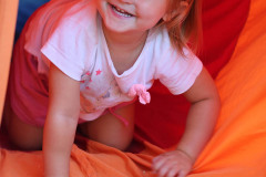 Uśmiechnięta dziewczynka na czworakach wychodzi z tunelu z pomarańczowego materiału.