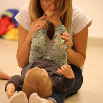 Kobieta siedzi z wyprostowanymi nogami, na których leży dziecko. Kobieta całuje gołe stopy dziecka.