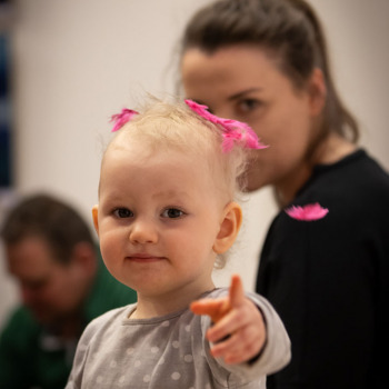 Zdjęcie portretowe dziecka z różowymi piórkami na głowie.  Patrzy w obiektyw i wskazuje palcem.
