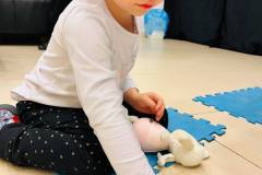 Dziewczynka klęczy na niebieskim puzzlu i przyciska dłonią leżący na podłodze woreczek z mazią.