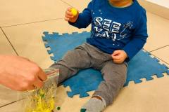 Dziecko siedzi na niebieskim puzzlu. W prawej dłoni trzyma kuleczkę. Przed nim ręka trzymająca woreczek z mazią.