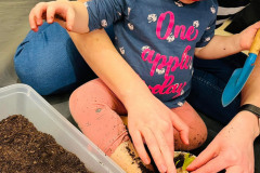 Dziecko siedzi przy plastikowym korytku z ziemią. Obejmują je ręce opiekunki, jednocześnie ugniatając ziemię w doniczce z cebulką.