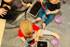 Zdjęcie robione z góry. Troje dzieci bawi się korytkami z ziemią. Łopatkami wsypują ziemię do kubków. Obok leżą różowe plastikowe miski.