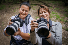 Dwie uśmiechnięte kobiety trzymają w dłoniach aparaty i kierują obiektywy w stronę fotografującego.