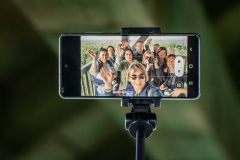 Grupa machających fotografów na wyświetlaczu telefonu. Telefon umieszczony na kijku do selfie.